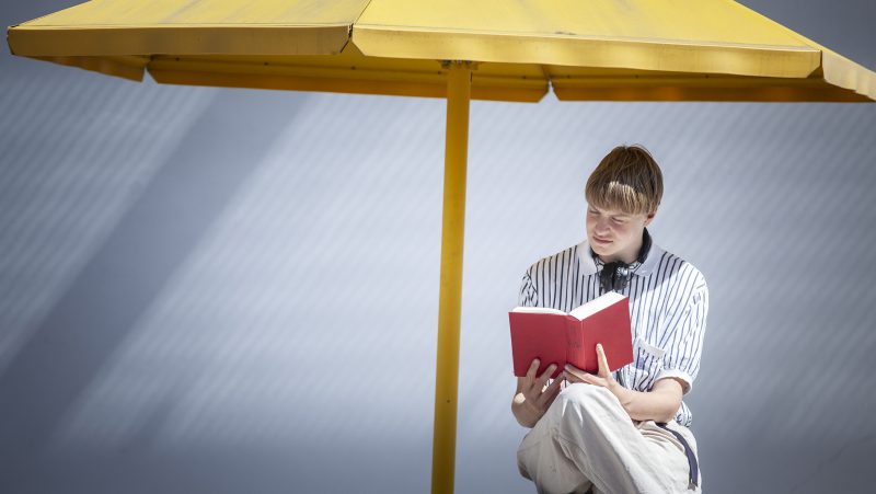 En kille sitter med en bok under ett stort paraply.